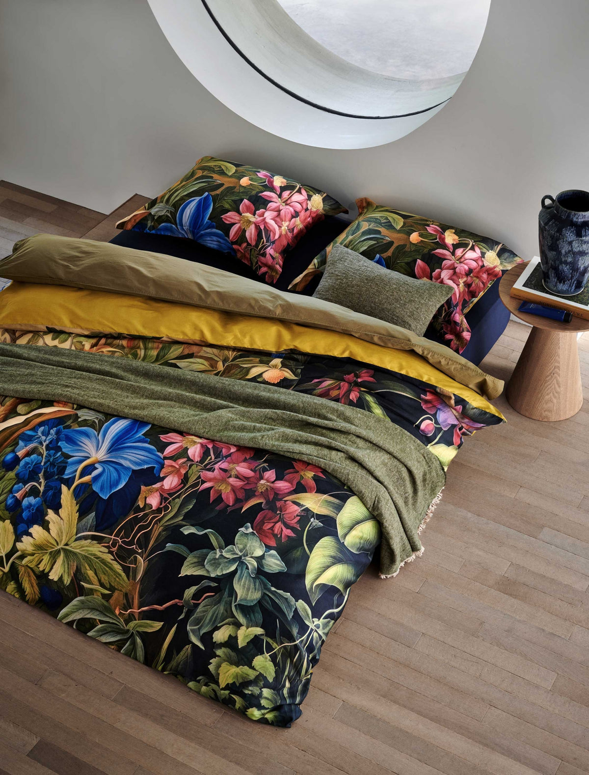 Bed in een rustige omgeving Mystic Garden multi dekbedovertrek heeft imposante bloemen in verschillende tinten roze en blauw, omgeven door grote bladeren en struiken. De achterkant van het dekbedovertrek is effen groen/oker