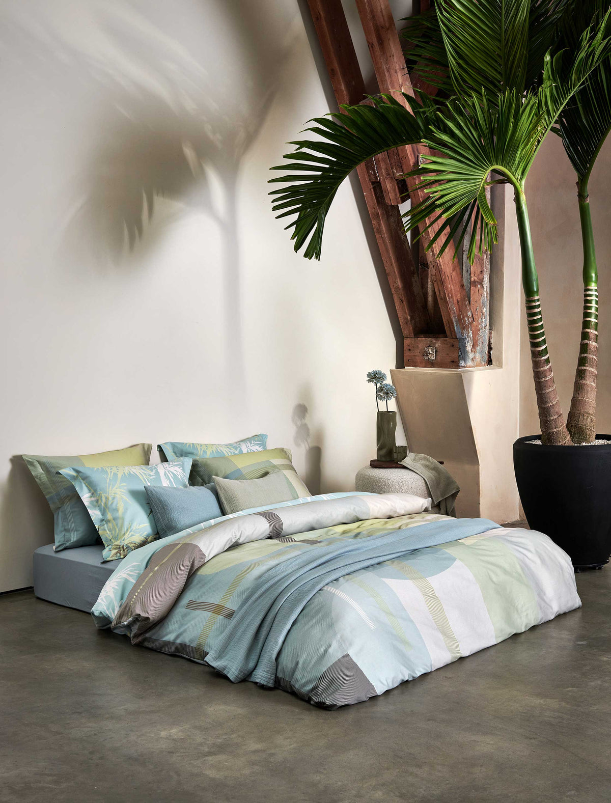 Lush Lagoon thyme dekbedovertrek heeft geometische vormen, waarbij strepen, rechthoeken en bogen in verschillende tinten groen, blauw en geel door elkaar heen lopen. De achterkant van het dekbedovertrek is effen lichtblauw. De kussens hebben aan beide zijden een paar kleurvlakken.